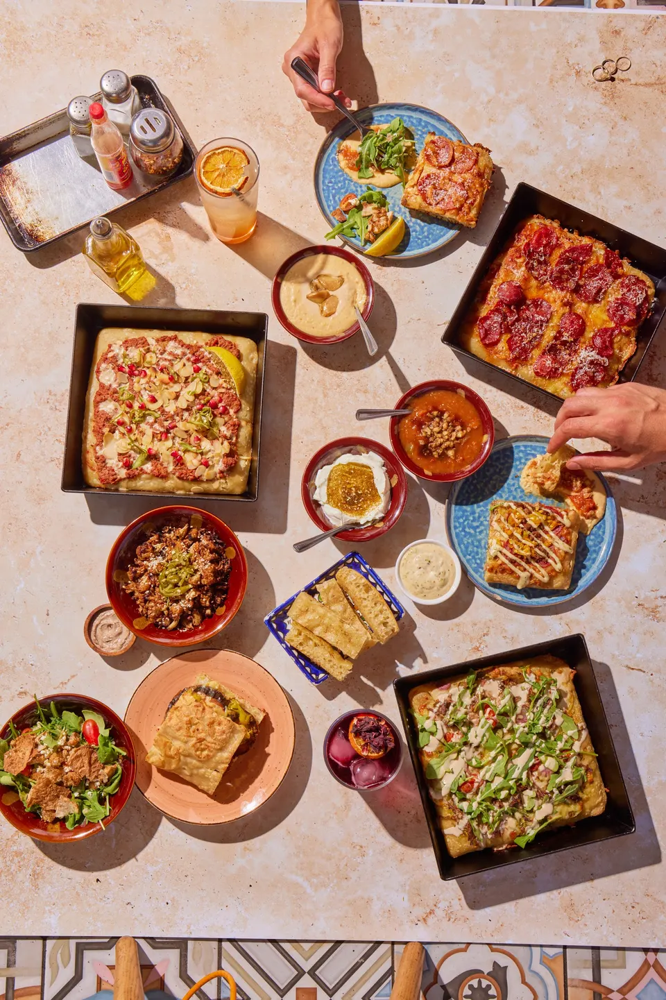 An overhead view of a table filled with various square pizzas, small bowls of dips, salads, and bread, with hands reaching in to eat.
