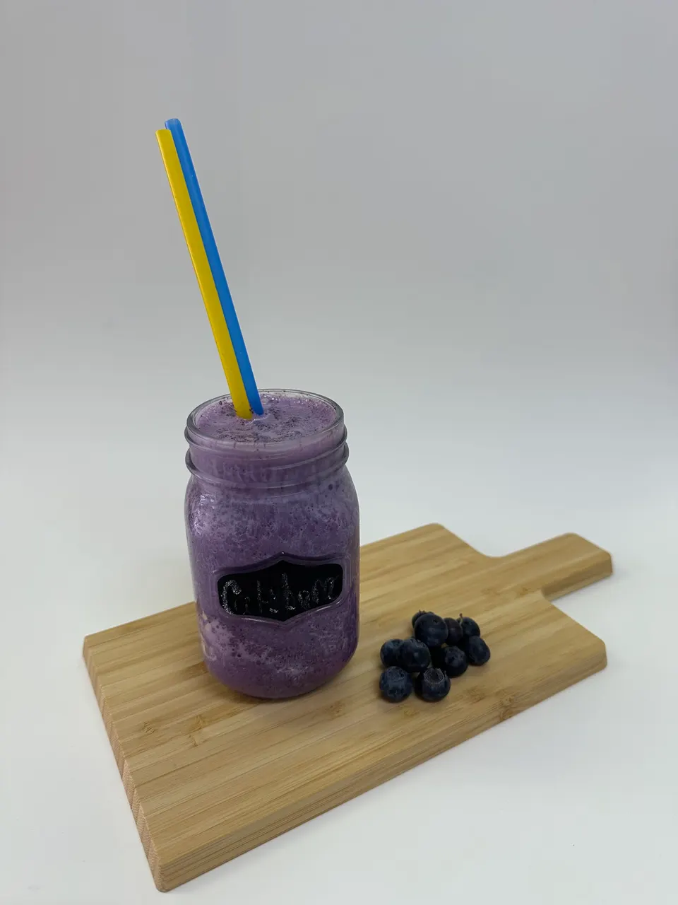 A purple blueberry smoothie in a glass jar with yellow and blue straws, served on a wooden cutting board next to a small pile of fresh blueberries against a plain white background.