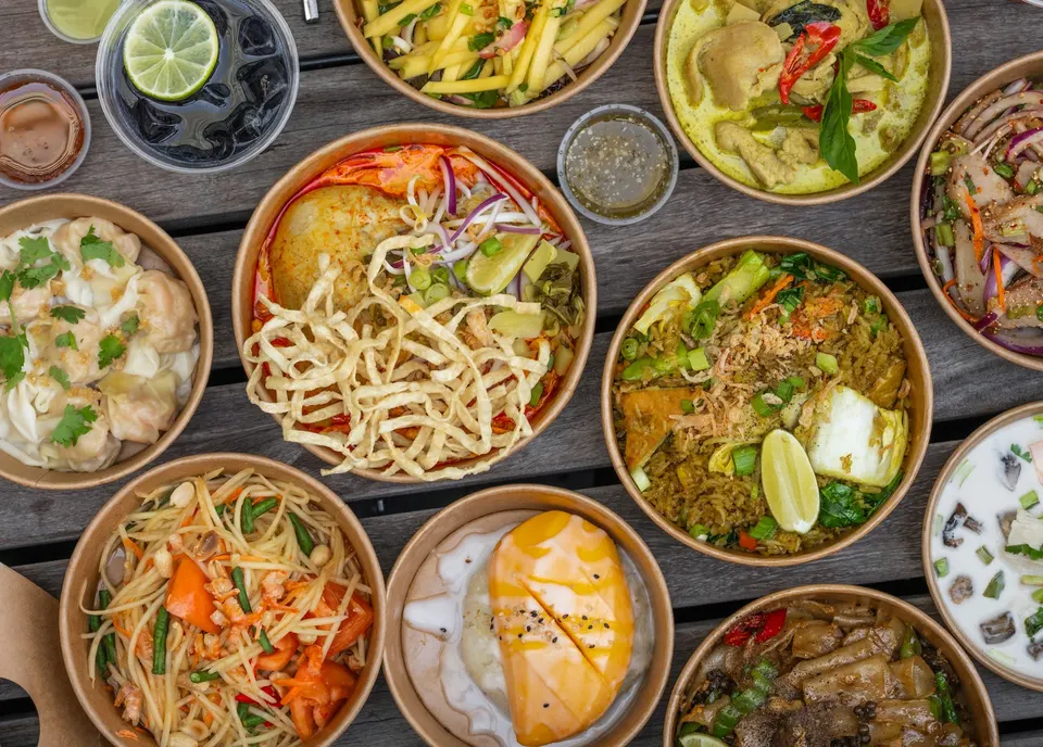 An overhead view of a wooden table filled with various Thai dishes in round paper bowls, including pad thai, green curry, mango sticky rice, and noodle soups.