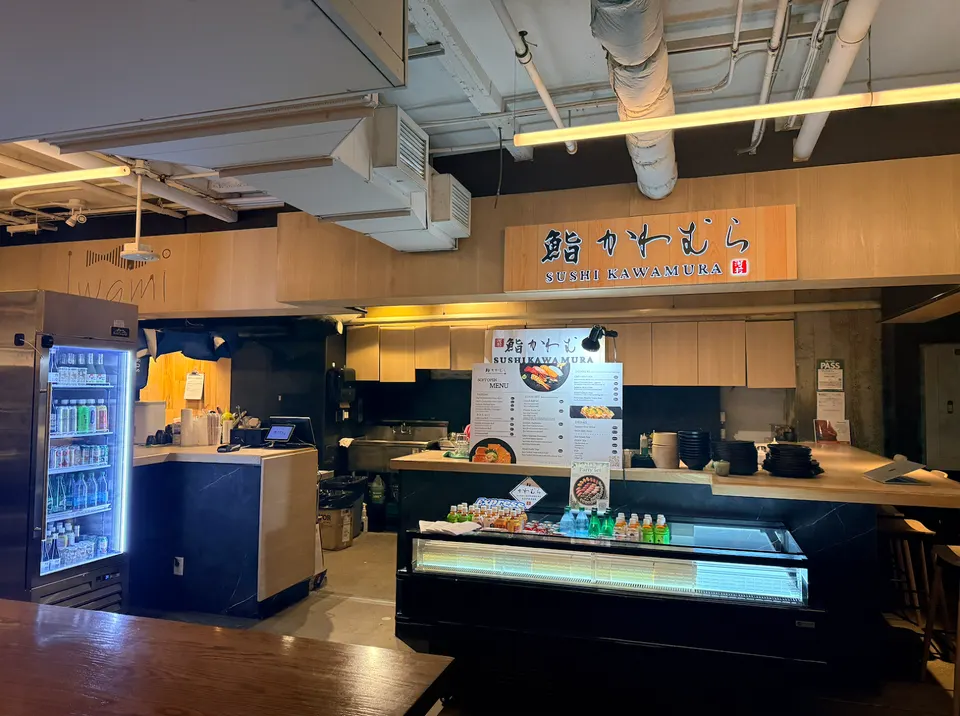 The counter and storefront of Sushi Kawamura inside a food hall, featuring a wooden sign with Japanese characters, a menu board, a refrigerated display case with drinks, and a beverage cooler on the left.