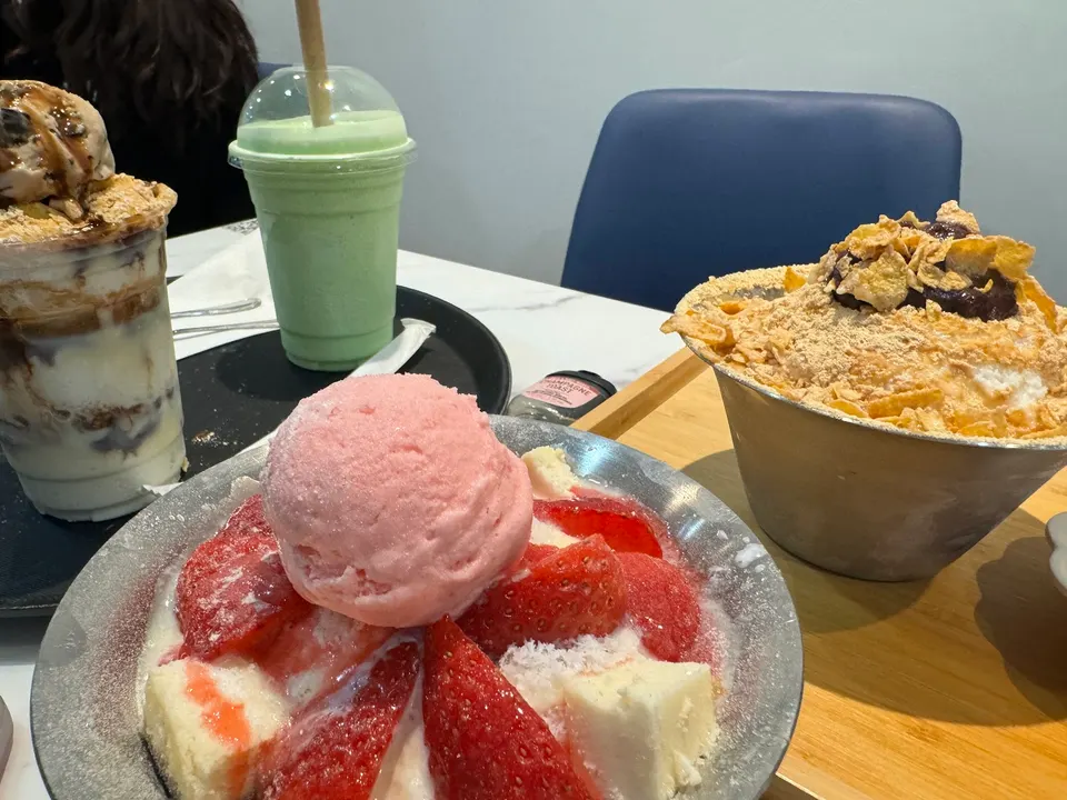 A table spread at HANBINGO featuring strawberry bingsu with pink ice cream, a traditional injeolmi bingsu with cornflakes, a green smoothie, and a layered dessert cup.