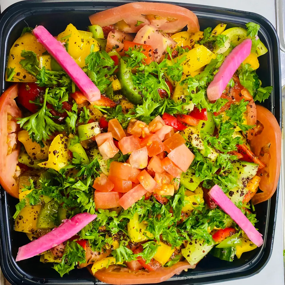 A vibrant, colorful salad in a black container, packed with chopped tomatoes, yellow peppers, green cucumbers, fresh parsley, and topped with bright pink pickled turnip spears.