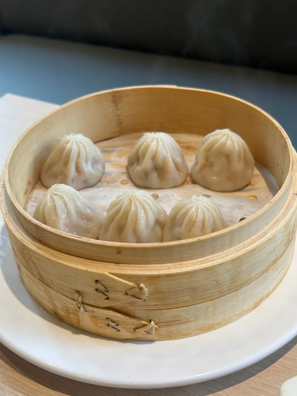 Six steamed soup dumplings, or xiaolongbao, arranged inside a round bamboo steamer basket placed on a white plate.