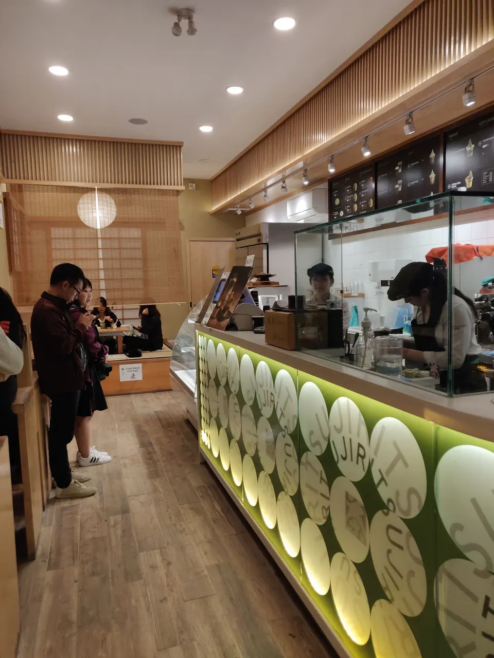 Interior view of Tsujiri Dundas showing customers waiting in line at a modern green and white counter while staff prepare orders behind a glass partition.