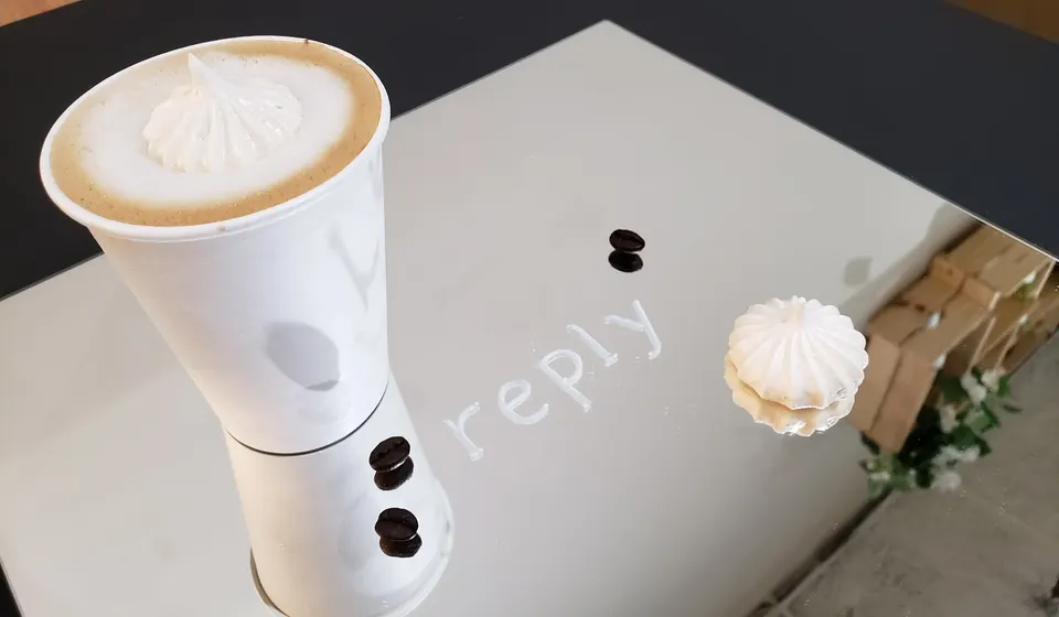 A white paper cup filled with a latte topped with a meringue cookie, placed on a reflective mirror surface next to a single meringue cookie, coffee beans, and the word 'reply' written on the glass.