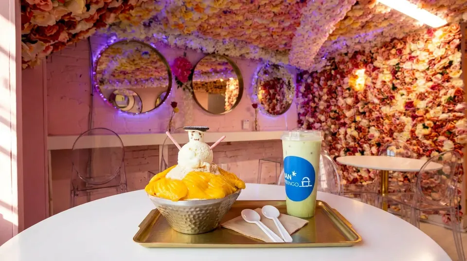 A mango bingsu topped with a snowman-shaped scoop of ice cream and a green iced drink served on a gold tray at a white table, set against a pink room decorated with floral walls and round mirrors.