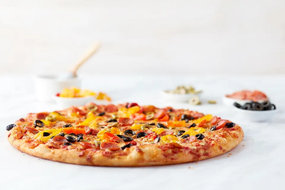 A freshly baked thin-crust pizza topped with melted cheese, pepperoni slices, black olives, and yellow and red bell peppers, set on a white surface with small bowls of ingredients in the background.