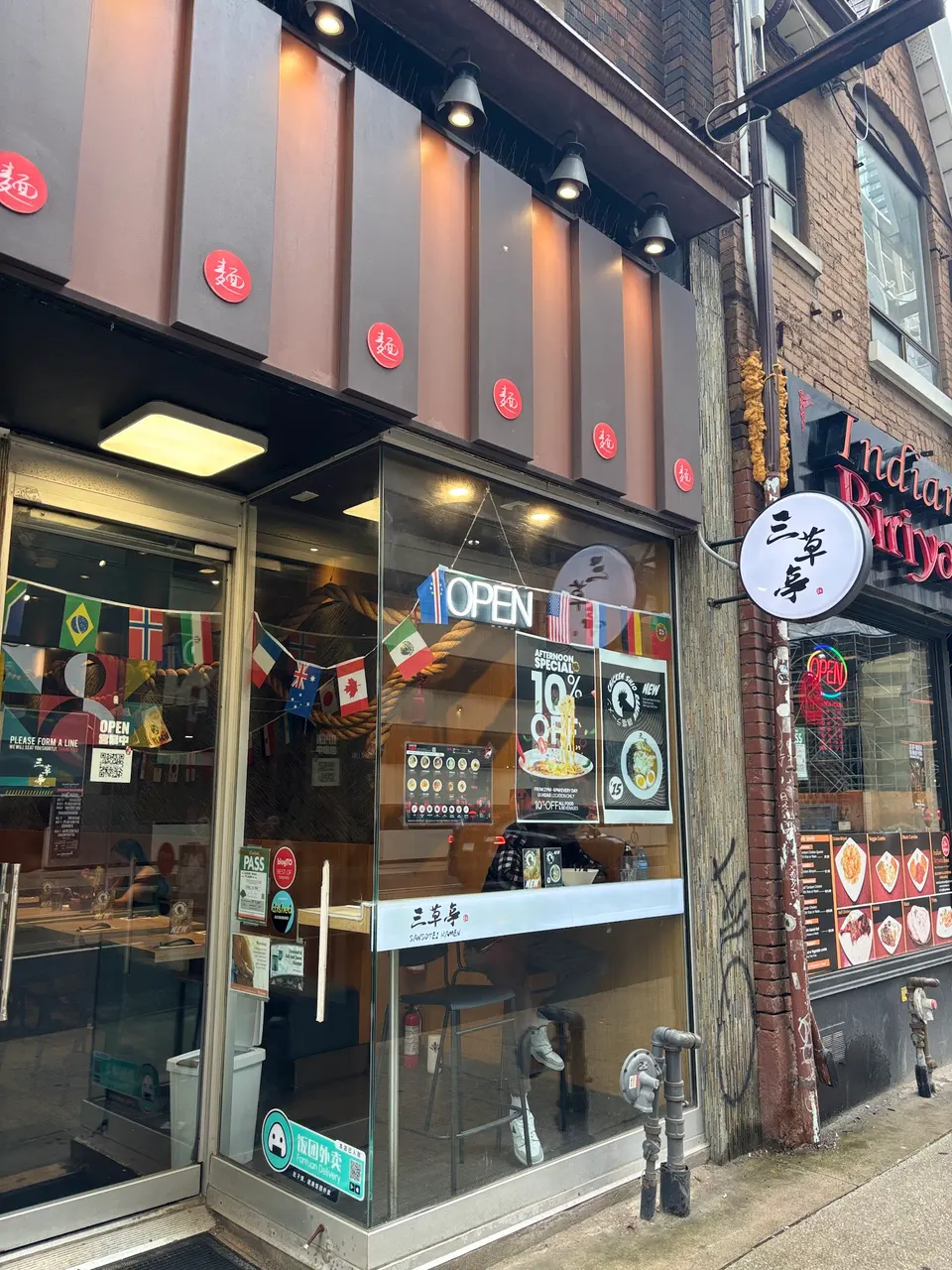 The storefront of Sansotei Ramen, featuring a glass window display with posters, hanging flags, and a round white sign with Japanese characters.