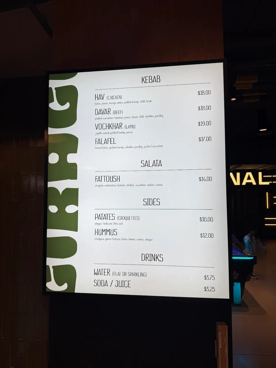 A brightly lit digital menu board displaying food and drink options with prices. The menu is divided into sections: Kebab, Salata, Sides, and Drinks. Dishes include Hav (Chicken), Davar (Beef), Vochkhar (Lamb), Falafel, Fattoush, Patates (Croquettes), and Hummus. Drinks listed are Water and Soda/Jui