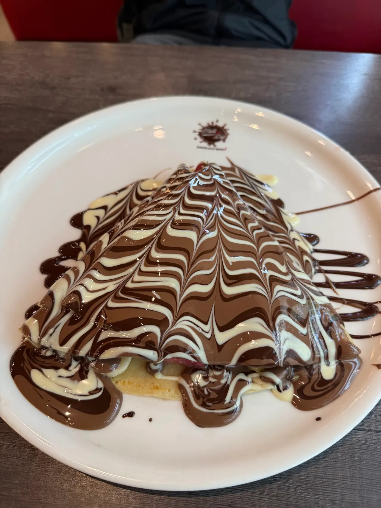 A triangular folded crepe on a white plate, lavishly drizzled with a marbled pattern of milk chocolate, dark chocolate, and white chocolate sauces.