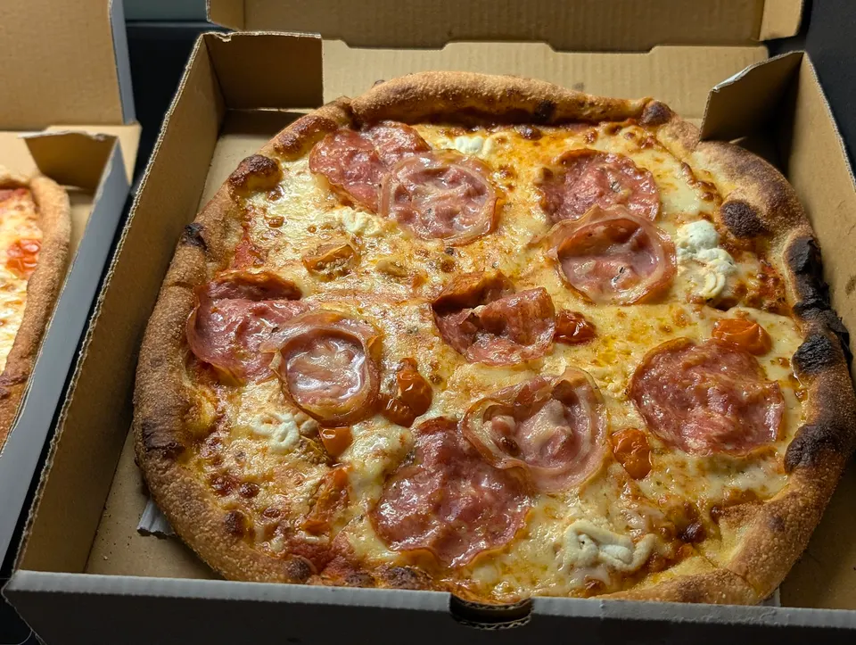 A freshly baked pizza with melted cheese, cured meats, and cherry tomatoes in a cardboard delivery box.