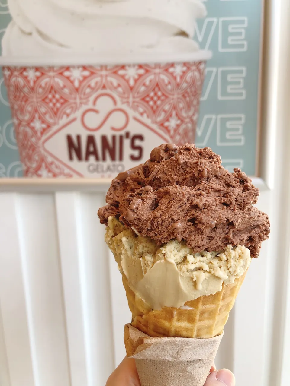 A hand holding a waffle cone with two scoops of gelato, a light brown scoop on the bottom and a dark chocolate scoop on top, in front of a Nani's Gelato sign.