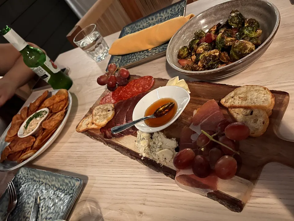 A wooden charcuterie board with cured meats, cheeses, grapes, honey, and sliced bread, alongside a bowl of roasted Brussels sprouts and a plate of chips on a light wooden table.