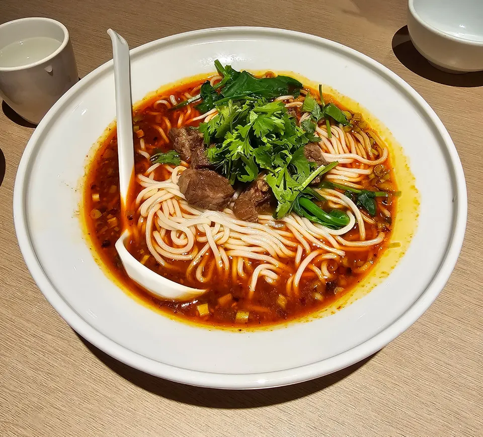 A large white bowl of spicy beef noodle soup with thick white noodles, chunks of beef, fresh cilantro, and green vegetables in a rich red broth, served with a white spoon.