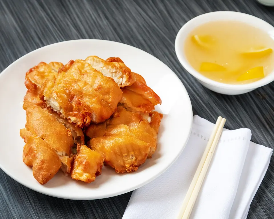 Crispy golden-brown battered chicken pieces served on a white plate next to a small bowl of yellow dipping sauce with lemon slices, chopsticks, and a white napkin on a dark wood table.