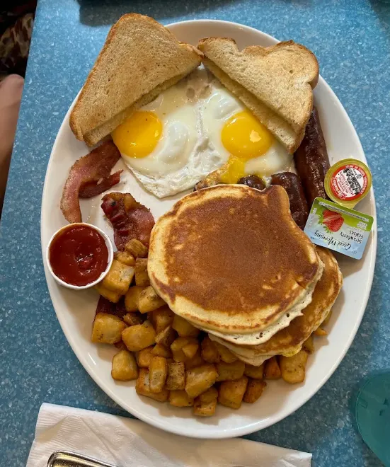 A full breakfast platter at Fran's Restaurant featuring two sunny-side-up eggs, toast, pancakes, home fries, bacon, sausage, ketchup, and jam packets.
