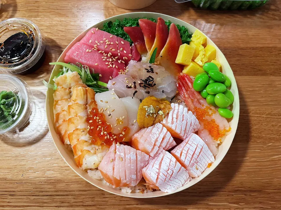 A colorful chirashi don bowl filled with assorted sashimi including salmon, tuna, shrimp, uni, and scallops, served with edamame and tamago.