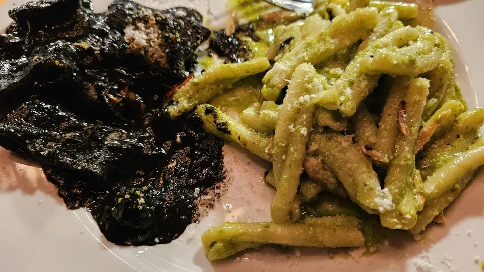 A plate of pasta split into two portions: one side features dark black squid ink pasta, and the other side features green pesto pasta sprinkled with grated cheese.