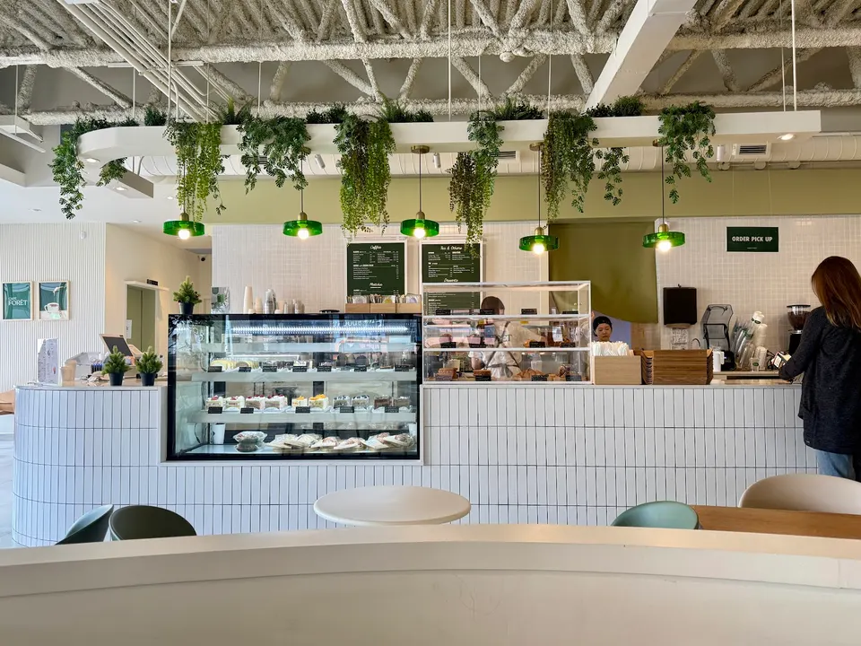A modern cafe counter with white vertical tiling, a glass pastry display case, hanging green pendant lights, and lush artificial plants draped from an overhead structure.