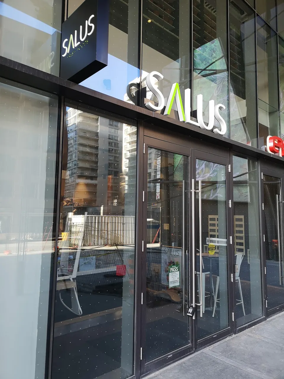 The glass storefront of Salus Fresh Foods, featuring a large white and green 'SALUS' sign above double glass doors reflecting nearby city buildings.