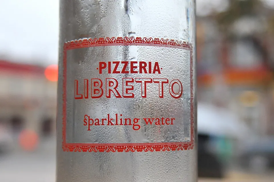 A close-up shot of a condensation-covered glass bottle featuring the red Pizzeria Libretto logo and the words 'sparkling water' printed on it, with a blurred street background.