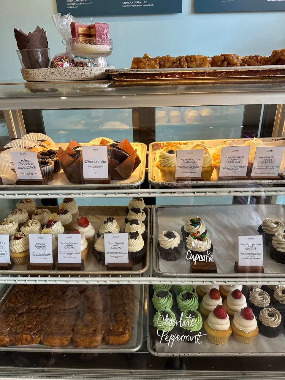 A bakery display case filled with gluten-free and dairy-free baked goods, including cupcakes, whoopie pies, lava cakes, and cinnamon buns, labeled with small signs.