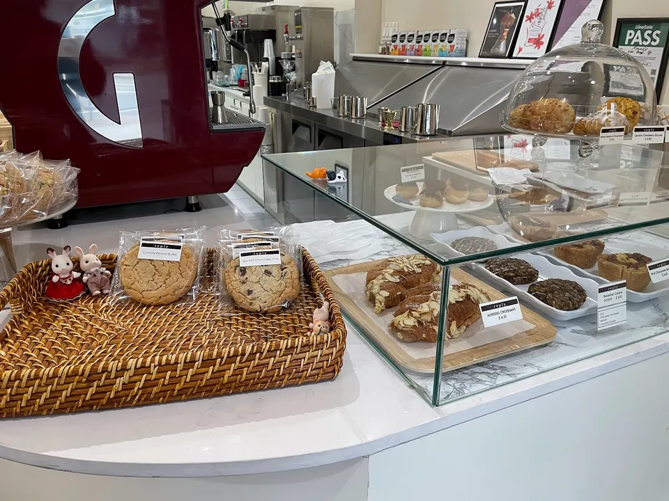 A bakery counter at Reply featuring a woven basket of packaged cookies, a glass display case with almond croissants, cookies, and pastries, and a red espresso machine in the background.