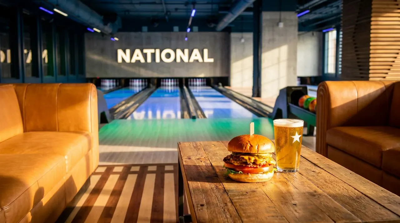 A juicy cheeseburger and a pint of beer with a star logo sit on a rustic wooden table, with leather couches and bowling lanes in the background under warm lighting.