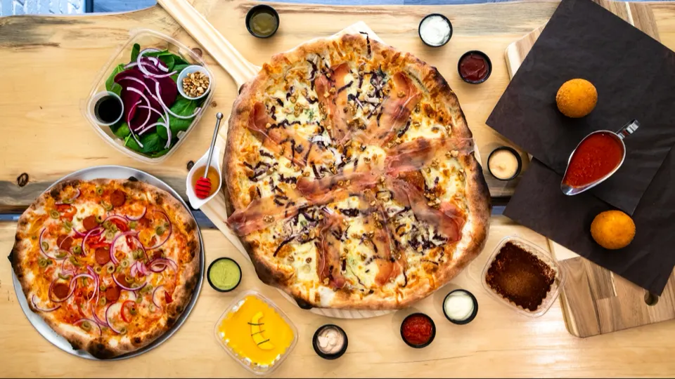 An overhead view of a wooden table filled with two pizzas, a spinach salad with red onions and beets, dipping sauces, arancini, and desserts.