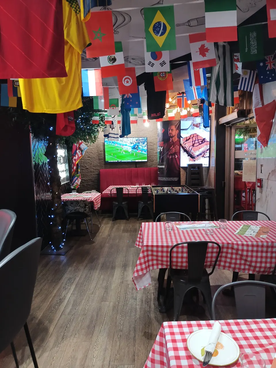 Interior of Pizza Rustica restaurant decorated with international flags hanging from the ceiling, featuring red and white checkered tablecloths, a foosball table, and a TV screen showing a soccer match.