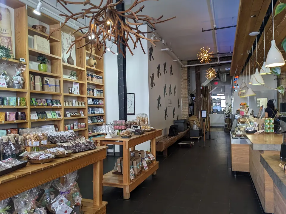 Interior of SOMA Chocolatemaker showing wooden shelves stocked with chocolate products, display tables with packaged treats, and a counter area under modern pendant lights.