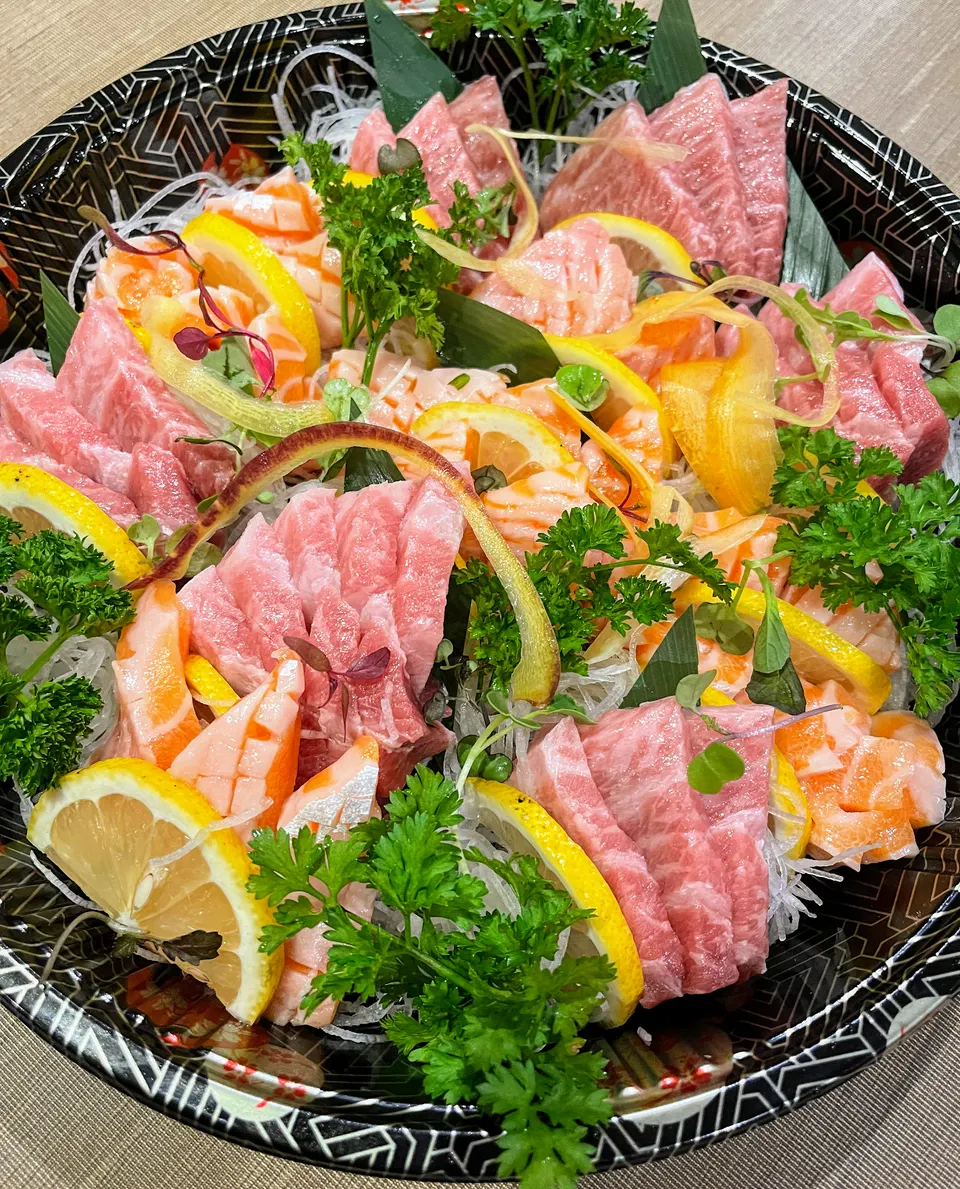 A large, colorful platter of fresh sashimi featuring thick cuts of pink tuna and orange salmon, garnished with lemon slices, parsley, and shredded white radish.