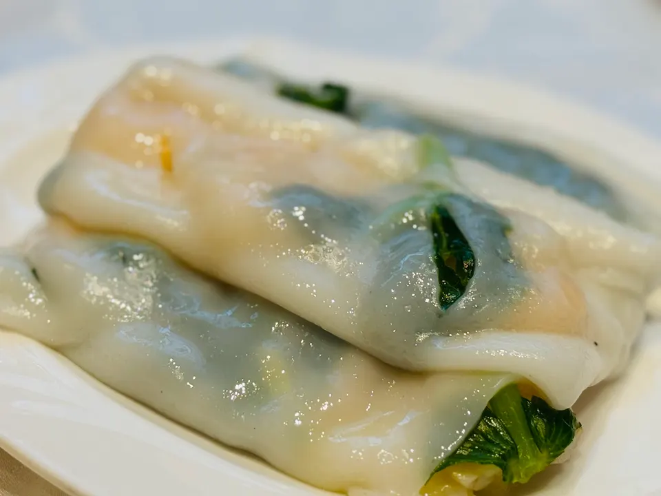 A close-up shot of steamed rice noodle rolls filled with shrimp and green vegetables, served on a white plate.