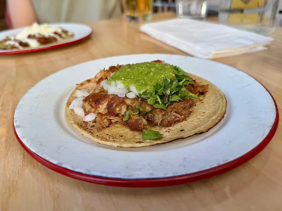 A single taco served on a white plate with a red rim, topped with shredded meat, chopped white onions, fresh cilantro, and green salsa.