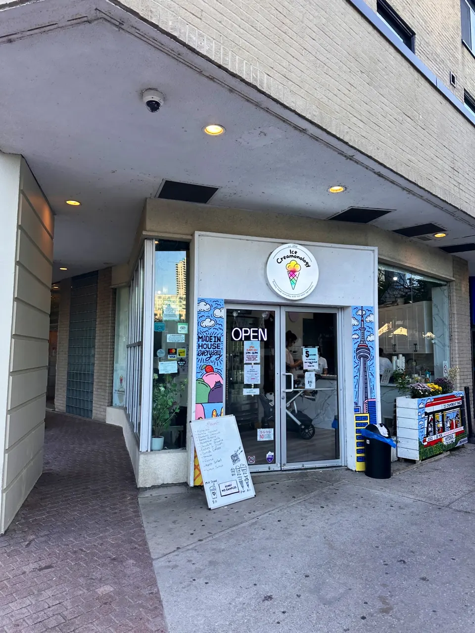 The storefront of Ice Creamonology features a glass double door with an 'OPEN' sign, colorful murals on either side, a white circular logo above, and a menu board on the sidewalk.