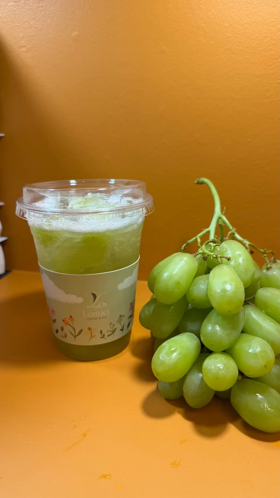 A clear plastic cup filled with an iced green grape drink next to a large bunch of fresh green grapes on an orange surface.
