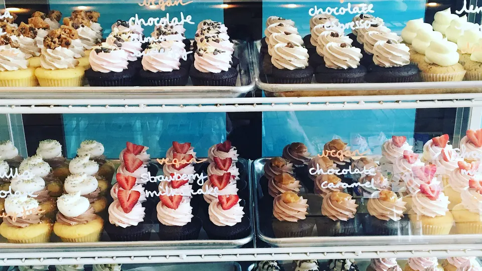 A display case filled with rows of beautifully decorated cupcakes, including chocolate with pink frosting, vanilla with cookie toppings, and strawberry-topped cupcakes.