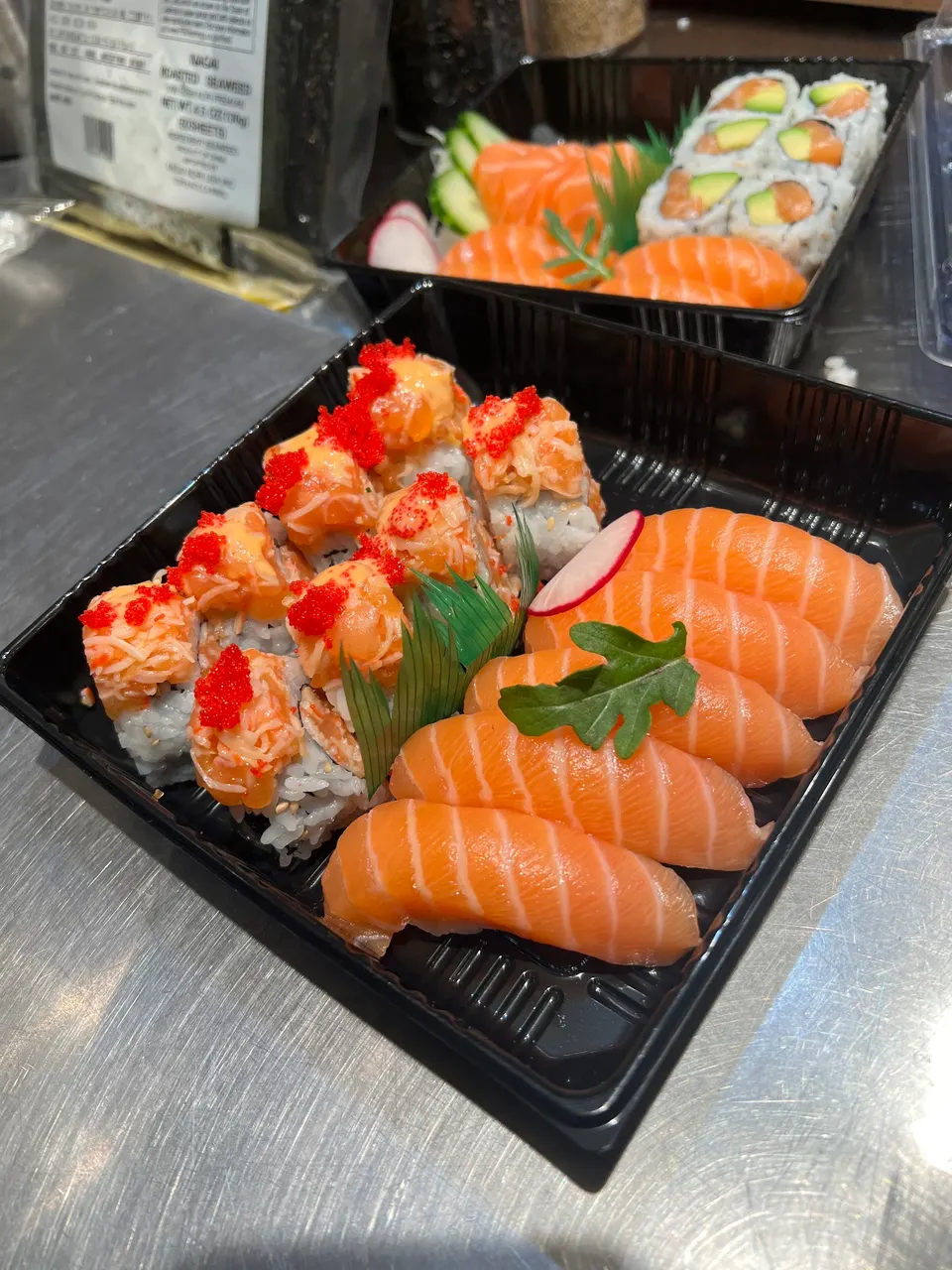 Two black plastic takeout containers filled with fresh sushi, featuring salmon nigiri and spicy salmon rolls topped with orange masago, resting on a stainless steel kitchen counter.