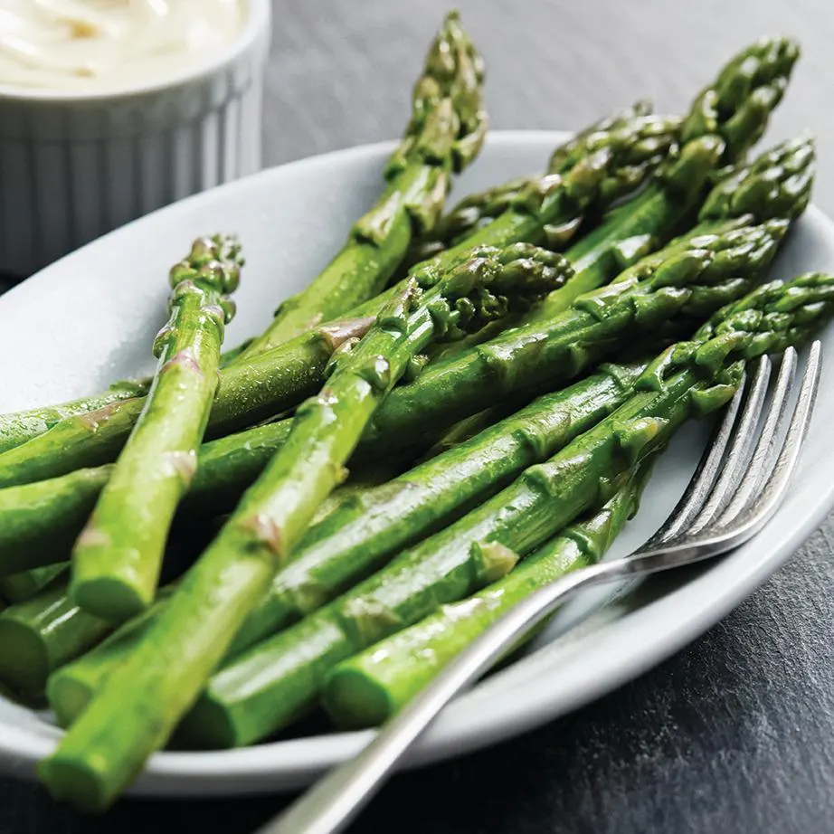 A close-up shot of freshly steamed green asparagus spears served on an oval white plate next to a silver fork, with a small ramekin of creamy sauce in the background.