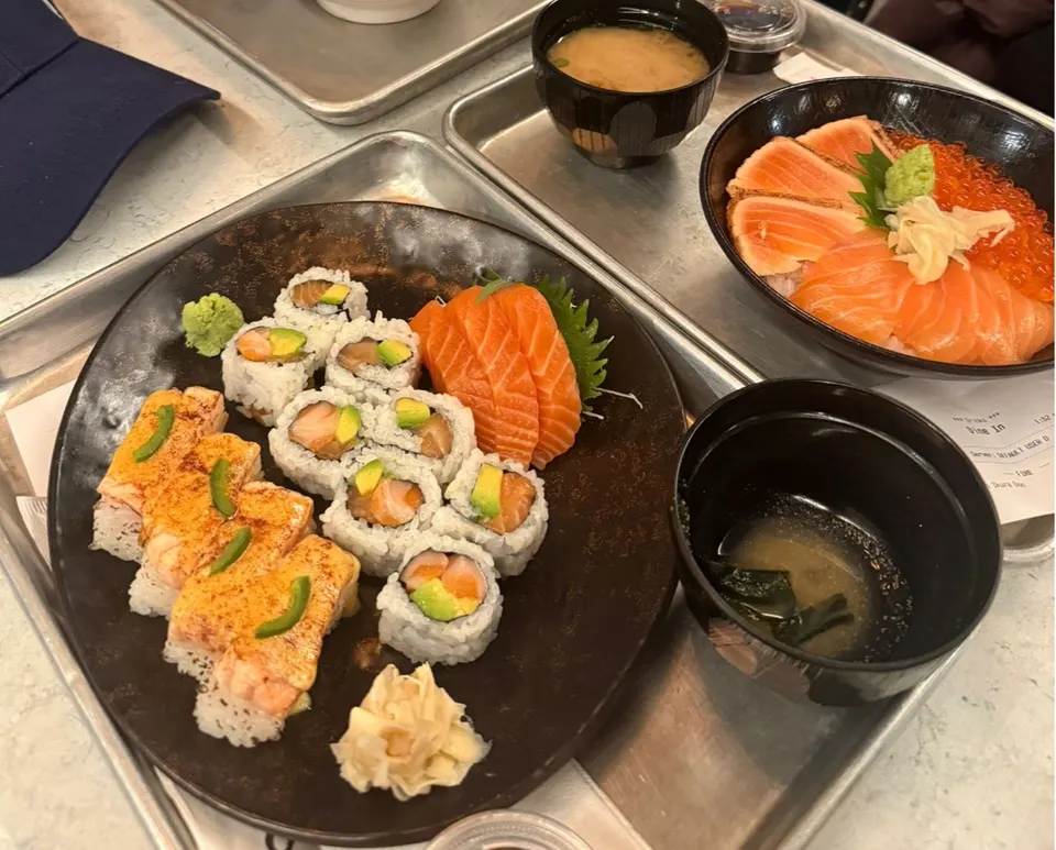 A metal tray holding a plate of sushi rolls and salmon sashimi, a bowl of salmon donburi with ikura, and two bowls of miso soup.