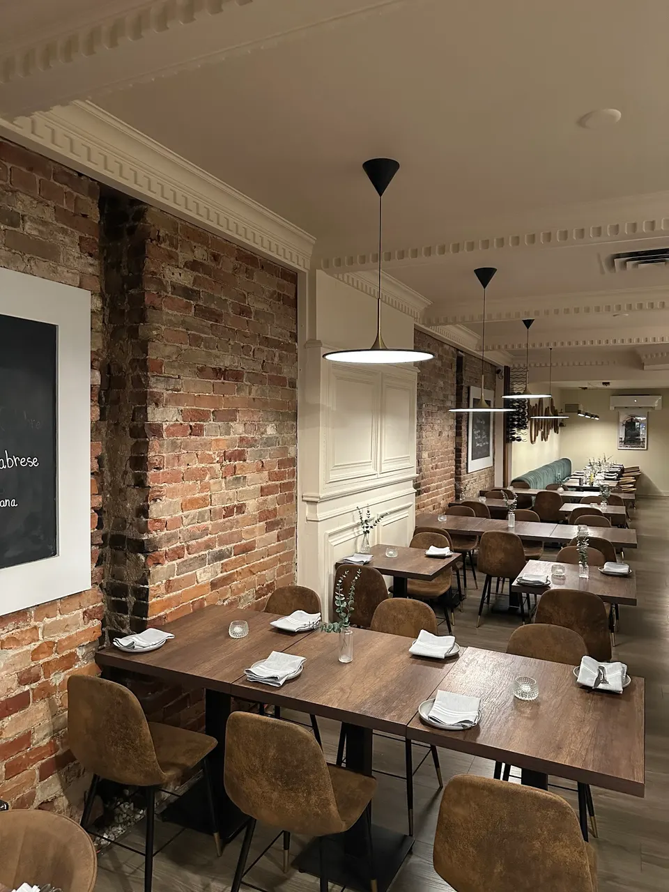 An empty dining room at Pizza e Pazzi featuring dark wood tables set with white napkins, brown upholstered chairs, exposed brick walls, and modern pendant lighting.