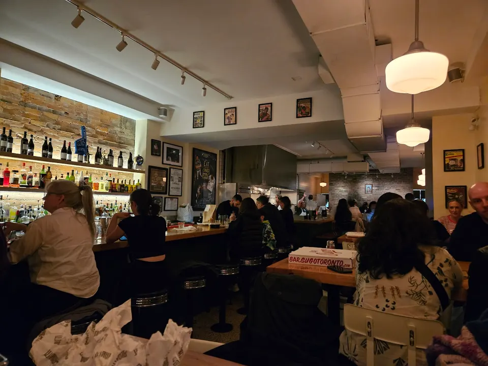 A cozy, dimly lit bar and dining area at Bar Sugo, featuring patrons seated along a wooden bar counter and at tables, with warm overhead lighting and a brick wall behind the bar shelves.
