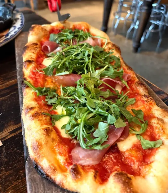 An oblong flatbread pizza topped with tomato sauce, melted cheese, prosciutto slices, and fresh green arugula, served on a wooden board.