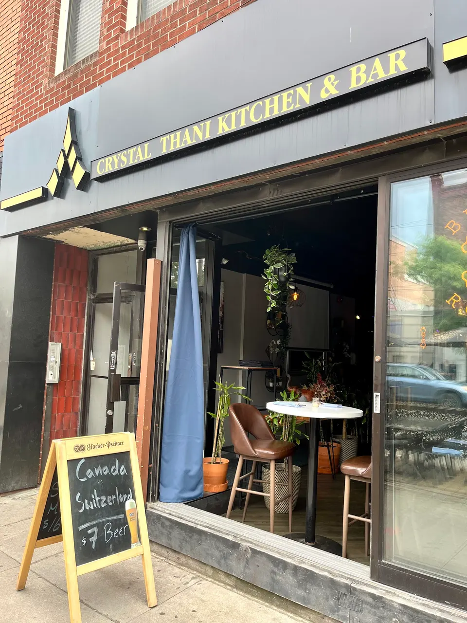 The storefront of Crystal Thani Kitchen & Bar, featuring a dark grey facade, a yellow sign, and an open glass door revealing a small dining area with high-top seating.