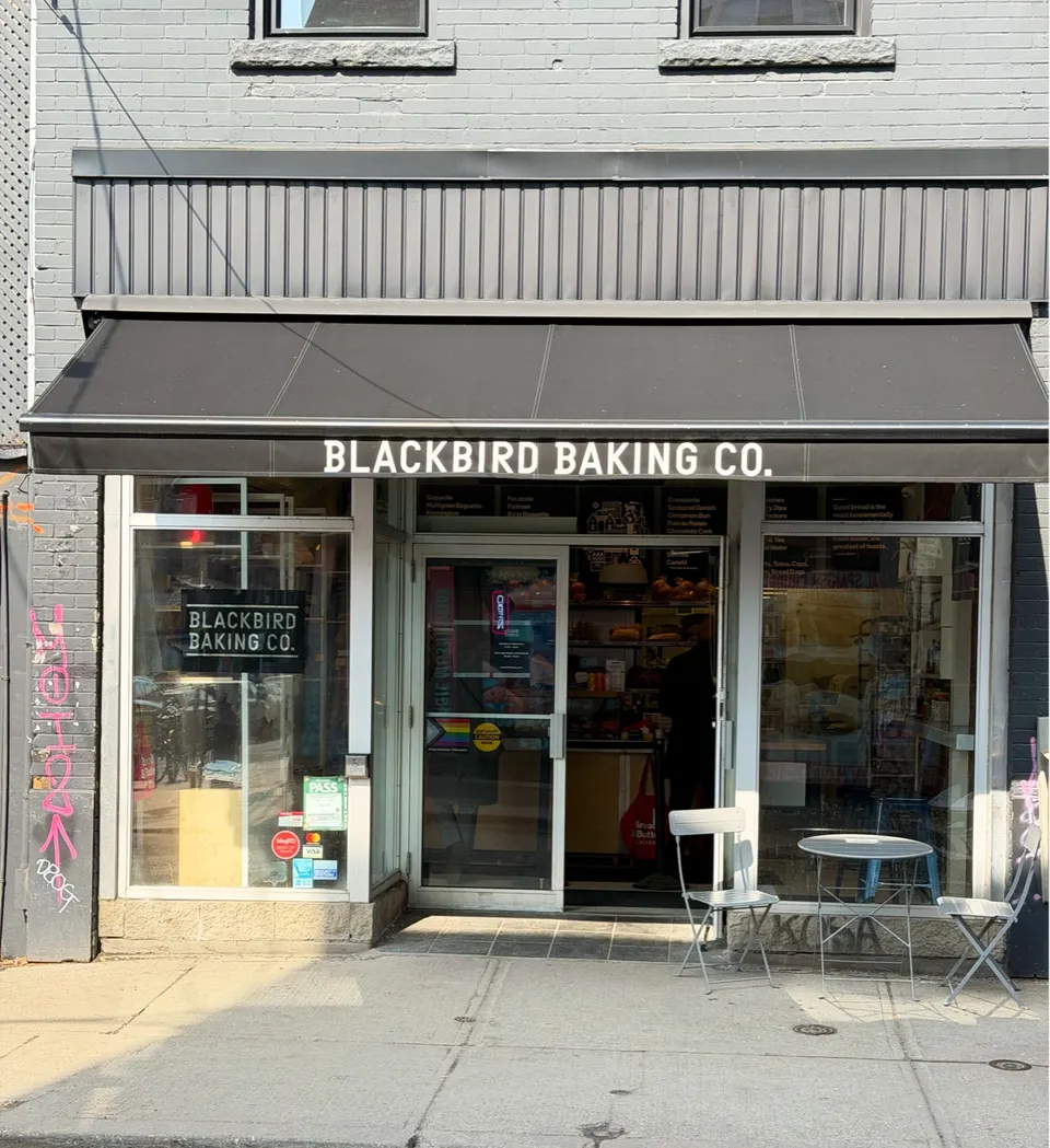 The storefront of Blackbird Baking Co. in Kensington Market, featuring a black awning with white lettering, large glass windows, and a small outdoor table with two chairs on the sidewalk.