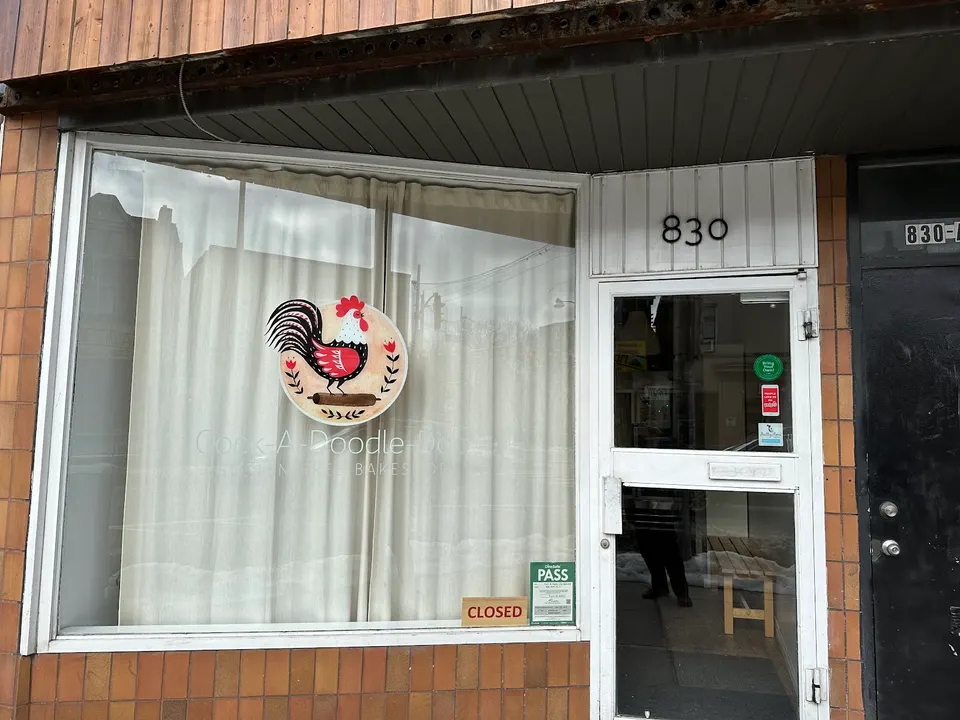Storefront of Cock-A-Doodle-Doo Gluten-Free Bakeshop featuring a large window with a rooster logo on a round sign, white curtains, and a glass door with the address 830 above it.