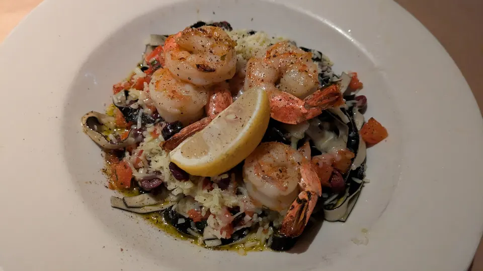 A plate of black squid ink pasta topped with grilled shrimp, diced tomatoes, olives, grated cheese, and a fresh lemon wedge in the center.