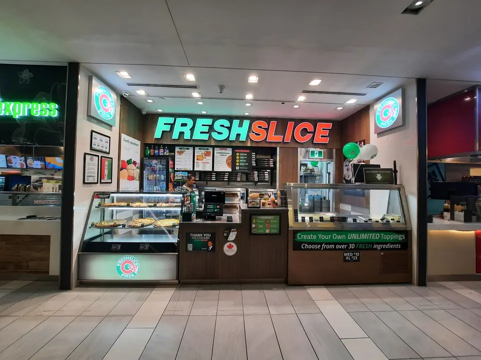A brightly lit Freshslice Pizza counter in a food court, featuring glass display cases with pizzas, a central ordering counter, and large green and orange signage above.