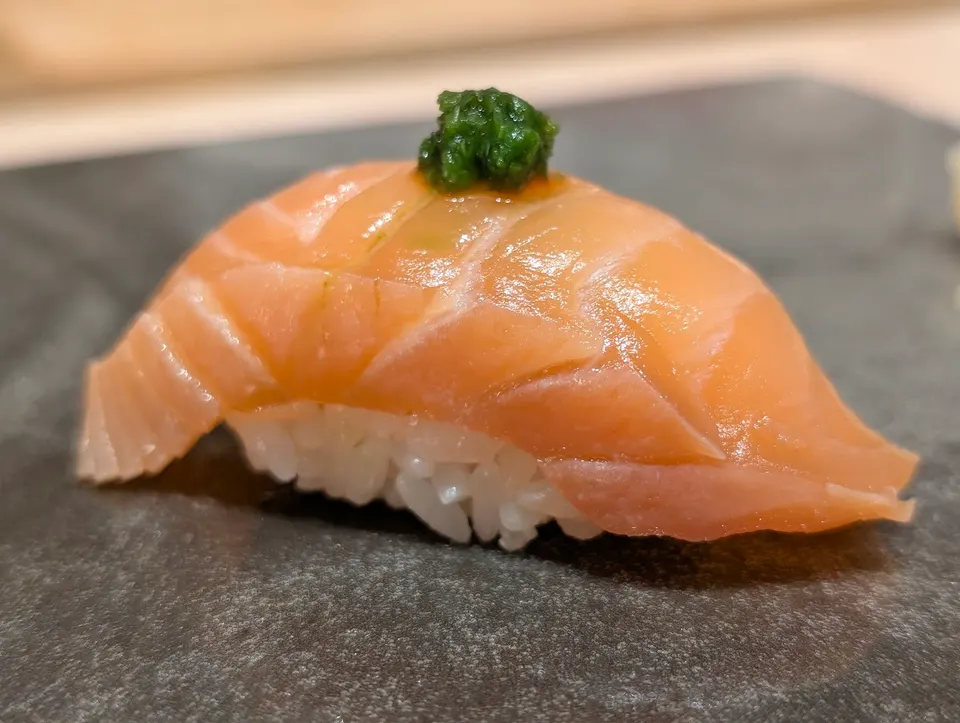 A single piece of salmon nigiri sushi topped with a small dollop of green herb paste, served on a dark textured slate plate.