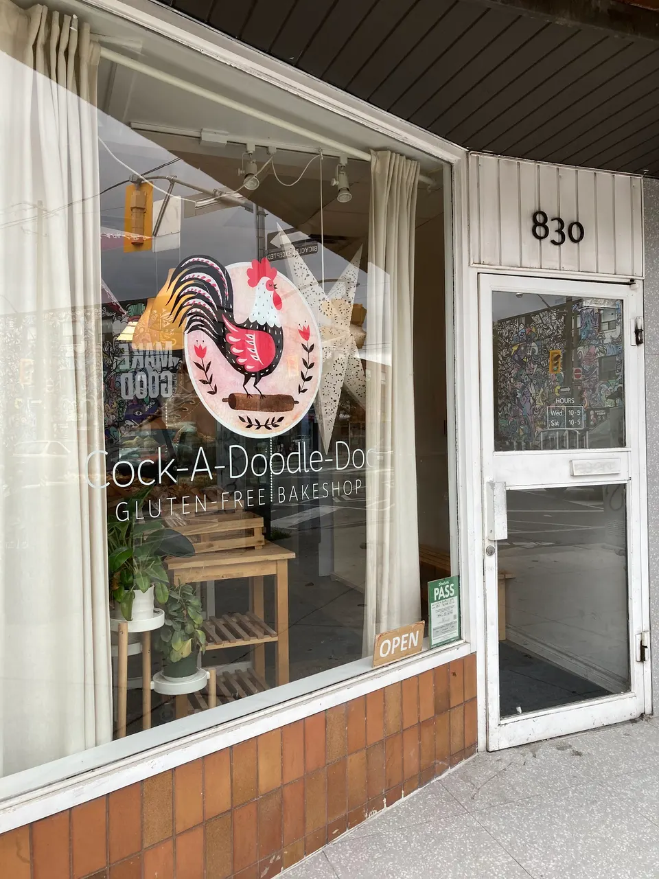The storefront of Cock-A-Doodle-Doo Gluten-Free Bakeshop, featuring a large window with a rooster logo, white curtains, and a white door with the address 830.